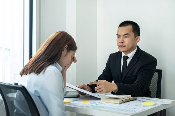 Businessman boss in suit scolding and pointing to woman office worker about bad work of her while she is sitting to stress and bow down in the office