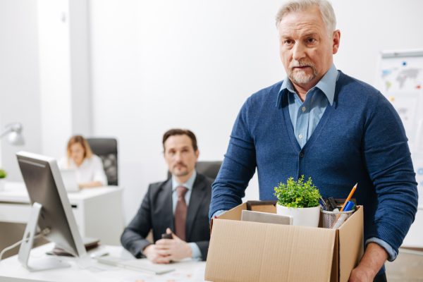 Quitting my responsibilities here. Fired involved aging employee standing and holding the box with his belongings while leaving the office