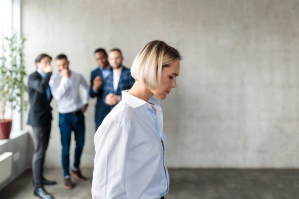 Workplace Bullying And Sexism. Male Colleagues Whispering Behind Back Of Unhappy Business Lady In Office. Victimization At Work. Female Discrimination Corporate Problem. Selective Focus, Panorama