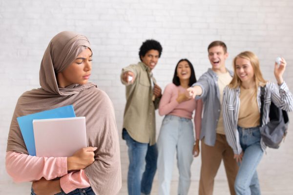 Upset lonely woman suffering from bullying and racial discrimination. Multiracial students throw paper at muslim lady with notebooks in college on brick white wall background , studio shot, free space