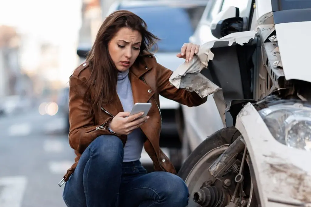 Driver checking her phone next to a damaged car after a Los Angeles car accident, highlighting the importance of documenting the crash and seeking legal help