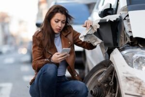 Driver checking her phone next to a damaged car after a Los Angeles car accident, highlighting the importance of documenting the crash and seeking legal help